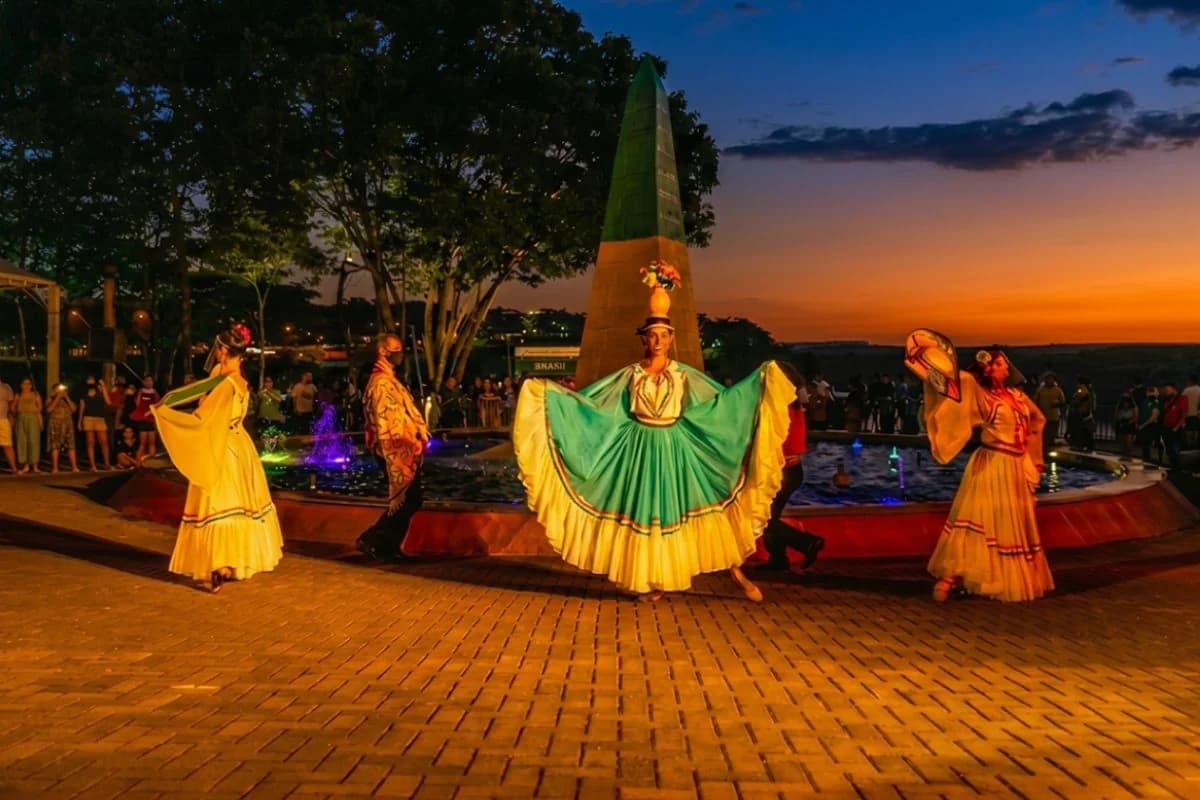 Show cultural no Marco das Três Fronteiras com dançarinas em trajes típicos paraguaios diante do obelisco brasileiro ao entardecer — Foto: Marco das Três Fronteiras / Grupo Cataratas