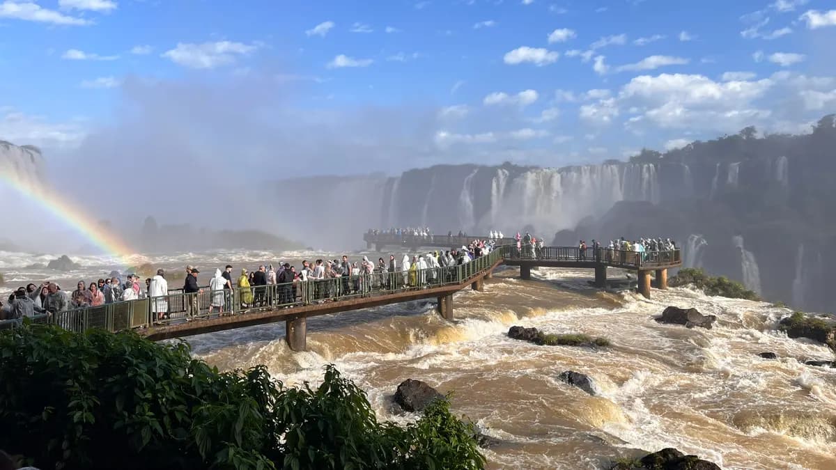 A passarela das Cataratas sobre o Rio Iguaçu barrento, com visitantes de capa de chuva e um arco-íris na névoa, no lado brasileiro